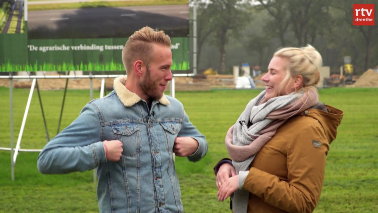Zwei Personen, ein Mann in einer Jeansjacke und eine Frau in einem braunen Mantel, stehen auf einer grünen Wiese und lachen miteinander, während im Hintergrund ein Transparent mit dem Schriftzug „De agrarische verbinding duizenden” zu sehen ist.