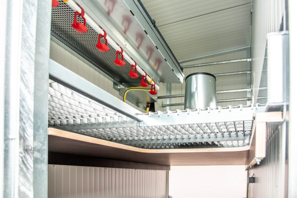 Innenansicht eines mobilen Hühnerstalls mit Metallregalen, roten Wasserspendern und einem silbernen Behälter.