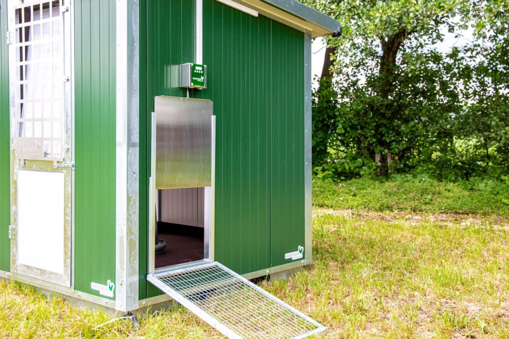Ein mobiler Hühnerstall in grüner Farbe mit einer geöffneten Tür und einer Rampe, um ins Innere zu gelangen, umgeben von Rasen und Bäumen.