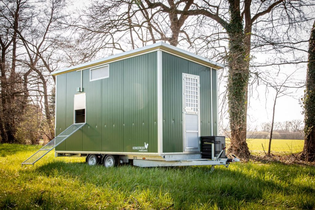 Mobiler Hühnerstall in grüner Farbe auf einem Anhänger, umgeben von Wiesen und Bäumen.
