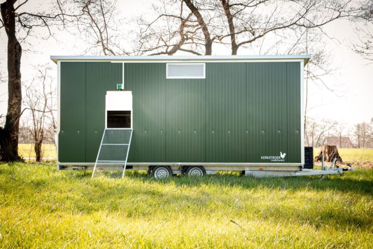 Ein mobiler Hühnerstall in Grün steht auf einem Wiesenstück, umgeben von Bäumen und frischem Gras.