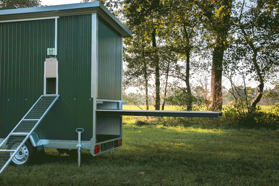 Eine mobile Hühnerstallanlage mit einer herausziehbaren Kotschublade und einer Rampensteighilfe, im Vordergrund eine Wiese und Bäume im Hintergrund.