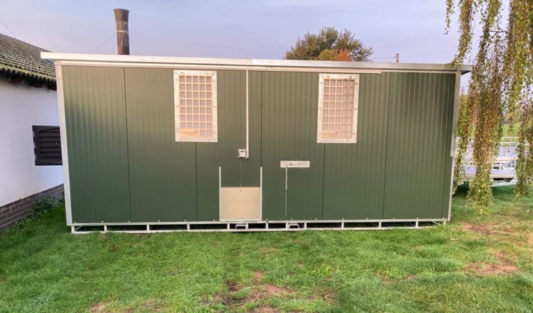 Ein mobiler Hühnerstall in Grün mit zwei Fenstern und einer seitlichen Tür auf einer Rasenfläche.