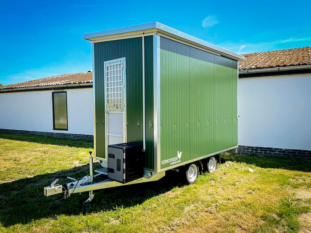 Ein mobiler Stall in grüner Farbe steht auf einem Grasfeld neben einem weißen Gebäude.