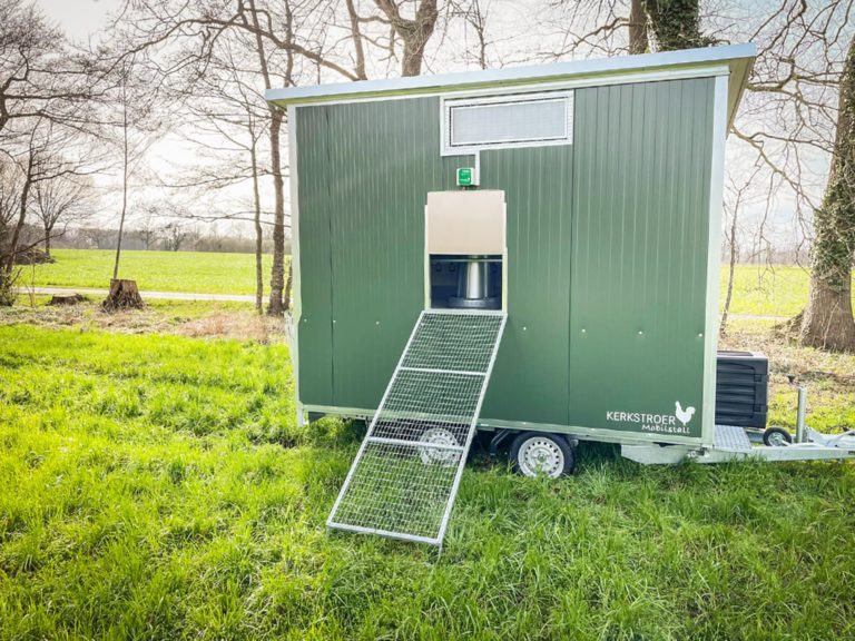 Ein mobiler Hühnerstall in grün mit Rampe steht auf einer Wiese, umgeben von Bäumen und einem offenen Feld.
