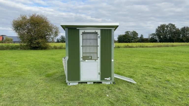 Ein mobiler Stall in grüner Farbe steht auf einer Wiese, umgeben von Bäumen und einem bewölkten Himmel.