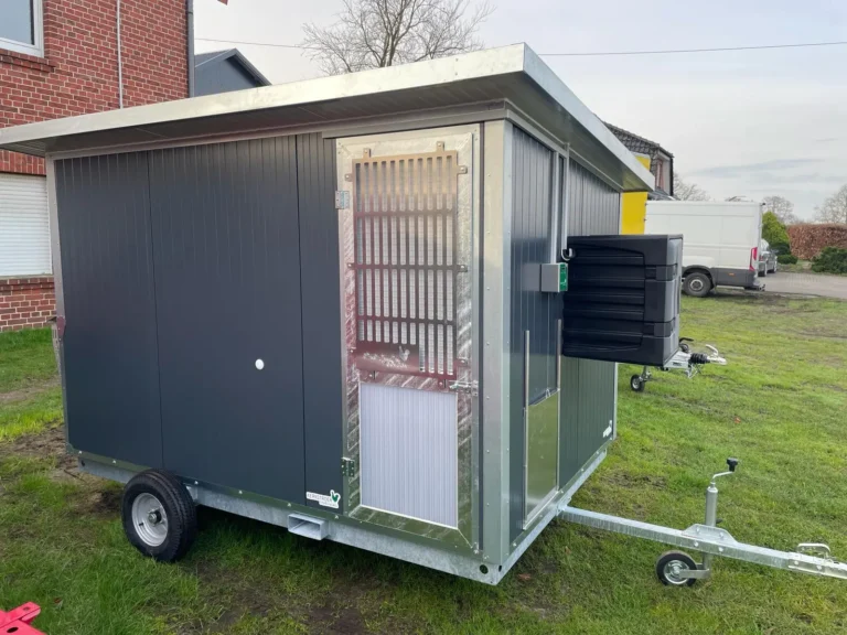 Ein grauer Anhänger mit einer Vorrichtung zum Ziehen, einer Tür und seitlichen Lüftungsschlitzen steht auf einem grünen Rasen.