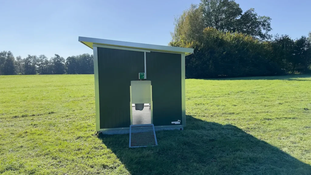 Ein mobiler Stall steht auf einer Wiese, umgeben von Bäumen und klarem Himmel.