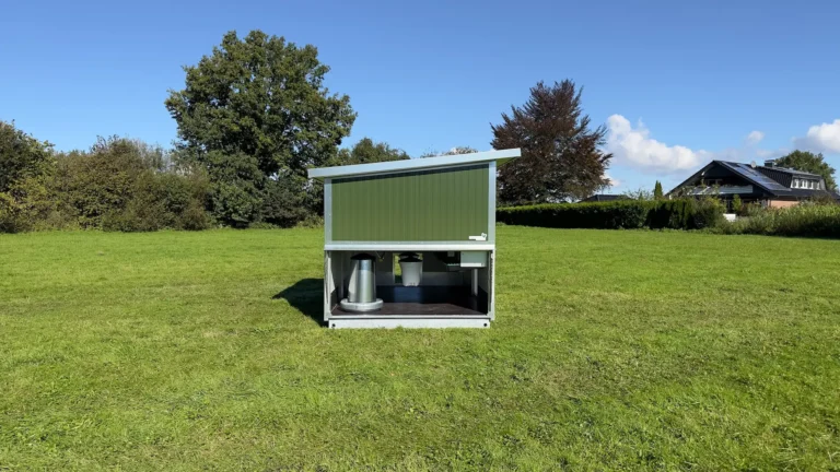 Ein mobiler Stall mit grünem Dach steht auf einer Wiese unter blauem Himmel, umgeben von Bäumen und einer Wohnhausfassade im Hintergrund.