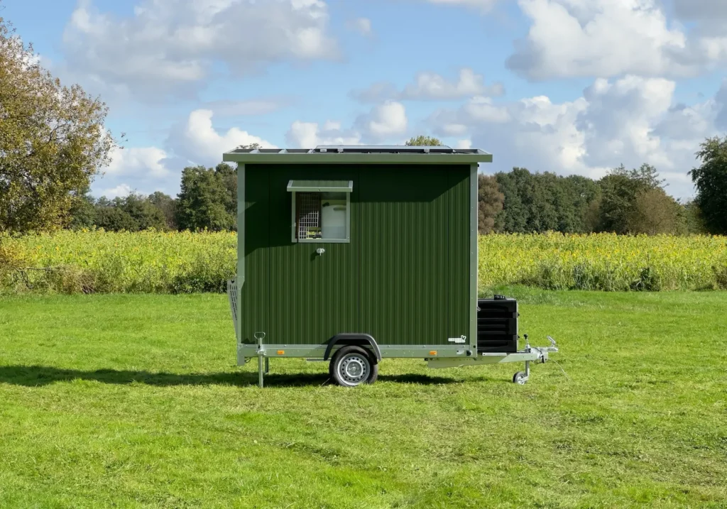 Ein grüner mobil Stall auf einem Feld, umgeben von Bäumen und einem blauen Himmel mit Wolken.